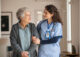 An Americare nurse in a blue dress talking to an older woman in a blue dress.