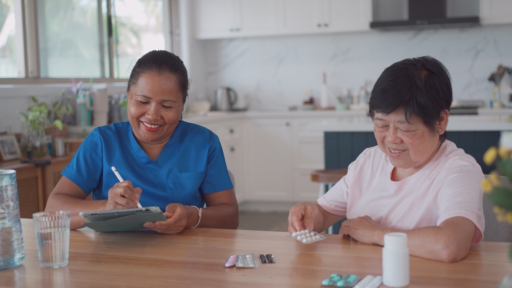An asian home care worker wearing blue scrubs and taking notes while an elderly patient is sorting medication sitting beside her. 
