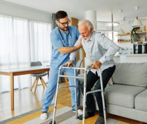 Male caregiver helping male senior patient stand up from the couch with his walker at home