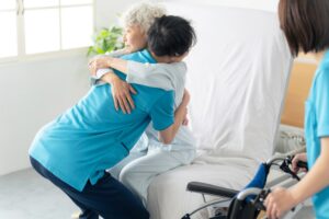 White haired Asian woman with arms around caregiver’s neck being lifted out of bed and into wheelchair.