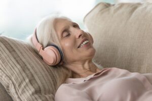 Grey-haired woman in pink blouse lying on sofa with eyes closed, listening to music on her pink earphones.