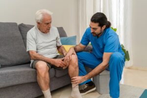 A male healthcare professional in blue scrubs examines the knee of an elderly man sitting on a couch, providing home-based care and support.