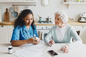 Personal care aide seated next to white-haired woman at a kitchen table reviewing medications.