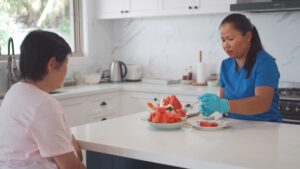 Asian home care nurse cutting and serving watermelon to an elderly Asian woman at her kitchen island.