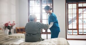 Elderly woman in striped sweater sitting on the edge of the bed being helped up by an asian caregiver dressed in scrubs.
