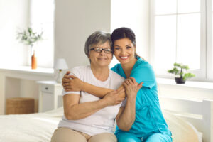 A young woman with dark hair, wearing blue scrubs, sits on a bed and embraces an older woman wearing a white t-shirt, glasses, and silver hair.
