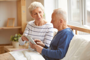 Older man in navy blue t-shirt propped up in bed reading something next to a white-haired woman wearing a white and blue striped top.