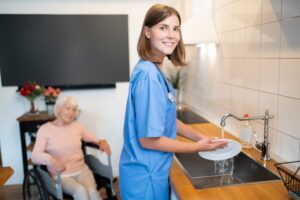 Short-haired care aide smiling while washing a dish at the kitchen sink, with an elderly client sitting in a wheelchair looking on in the background
