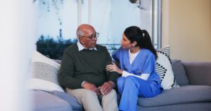 A south asian caregiver holding the arm and listening intently to a south asian man as he speaks while seated on the sofa.
