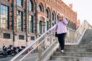  An older woman walking down a set of stairs outside wearing a purple blouse, navy pants, and eyeglasses.