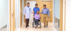 Asian woman wearing a purple top in a wheelchair, holding her husband’s hand, as she’s wheeled out of the hospital, accompanied by a doctor and nurse.

