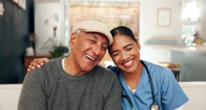Asian home health aide smiling and giving a hug to her male patient who is wearing a grey sweater and cap.