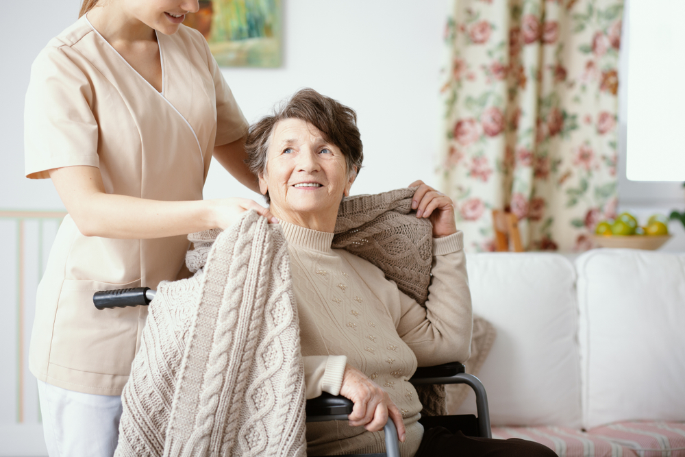 An elderly woman in a wheelchair is being covered with a blanket by a nurse dressed in beige scrubs.