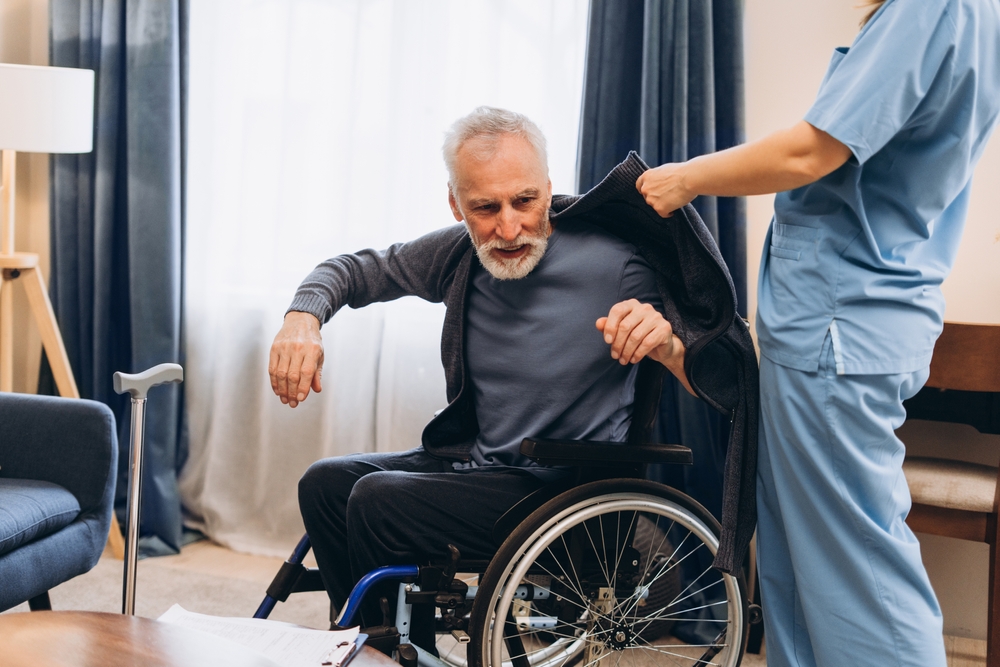 A man in a wheelchair getting help putting his coat on from a caregiver