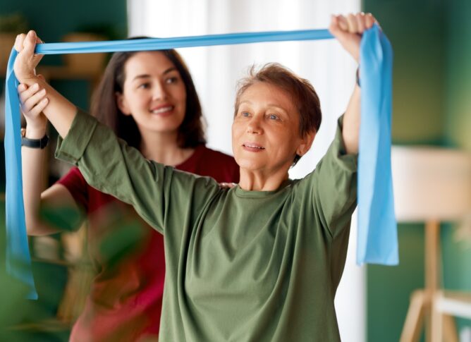An older woman in a long sleeve green shirt using a stretch band with the assistance of her in–home care giver standing behind her.