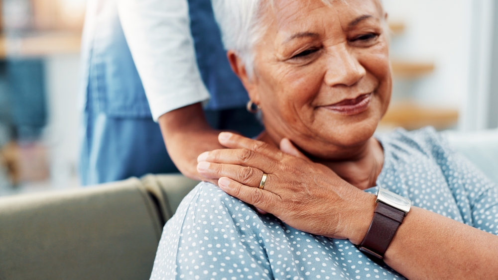 An elderly woman wearing a green and white polka-dotted top, patting a home care worker's hand that’s resting on her shoulder.