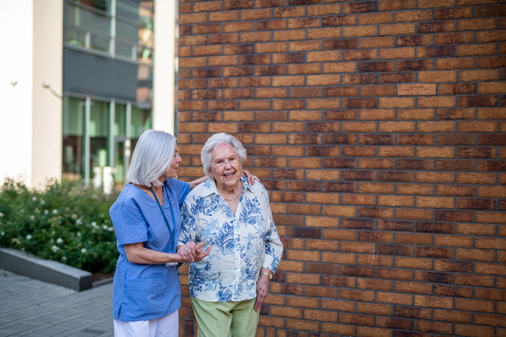 Woman with white hair, blue blouse, and green pants, walking with a female health aide wearing blue scrubs with silver hair.
