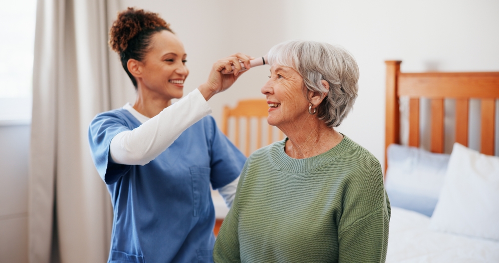 Silver-haired woman wearing a green sweater, having her hair combed by a home health worker wearing blue scrubs.