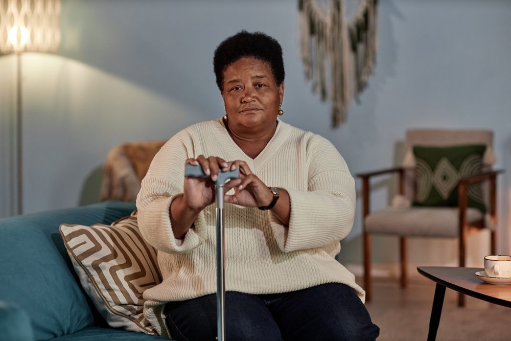 A black woman wearing a cream sweater holding onto her cane while sitting on a blue sofa.