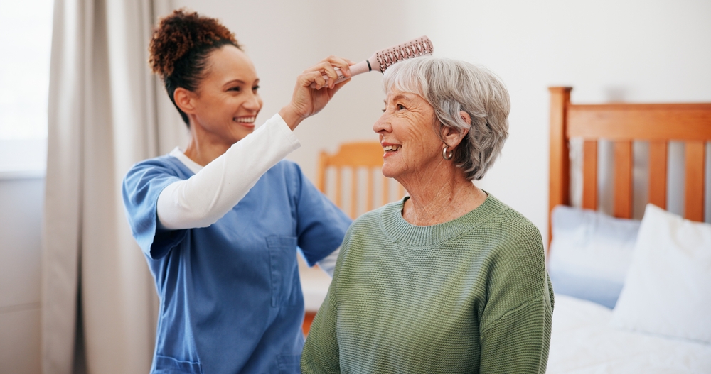 A woman with grey hair wearing a green sweater and having her hair brushed by a home care worker wearing blue scrubs while sitting on a bed.
