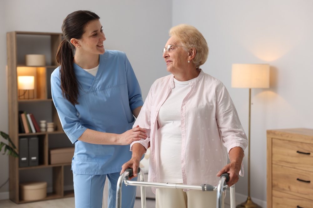 A white-haired woman wearing a pink blouse and using a walker, being assisted by a brown-haired ponytailed home care worker wearing scrubs. 