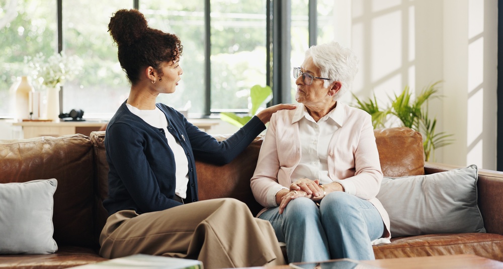 A senior woman with glasses and short silver hair sitting with a caregiver who’s resting a hand on her shoulder.
