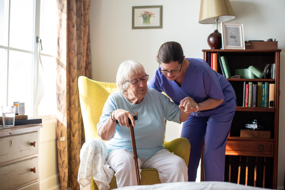 White-haired woman with glasses and a cane being assisted out of her chair by a caregiver in blue scrubs and glasses.