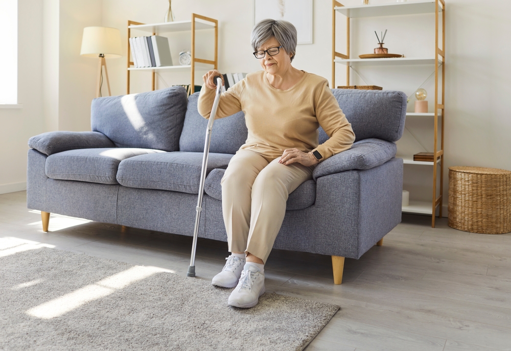 An older woman wearing beige clothing and using a cane to stand up from her sofa.