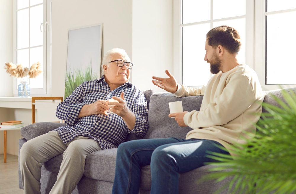 A father wearing a checked shirt and glasses speaking with his son while sitting on the sofa.