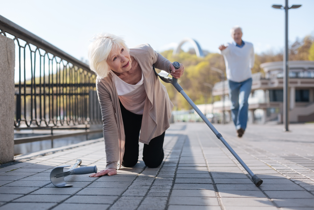 A white-haired woman recovering from a fall on the pavement using a pair of elbow-supported walking canes, with a man running to her aid in the background.