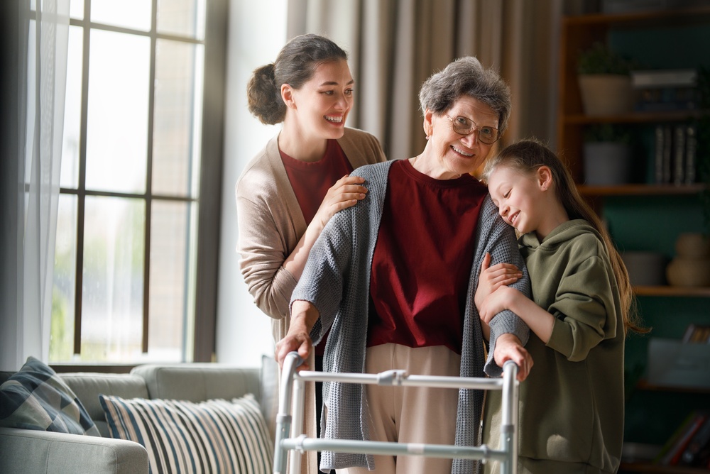 A senior woman using a walker is being hugged by her young granddaughter, while another woman smiles at her over her right shoulder.