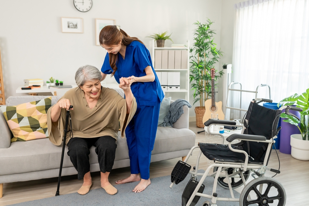 An older woman with a cane being assisted while getting up from the sofa by a home care professional wearing blue scrubs and helping her sit in a wheelchair.