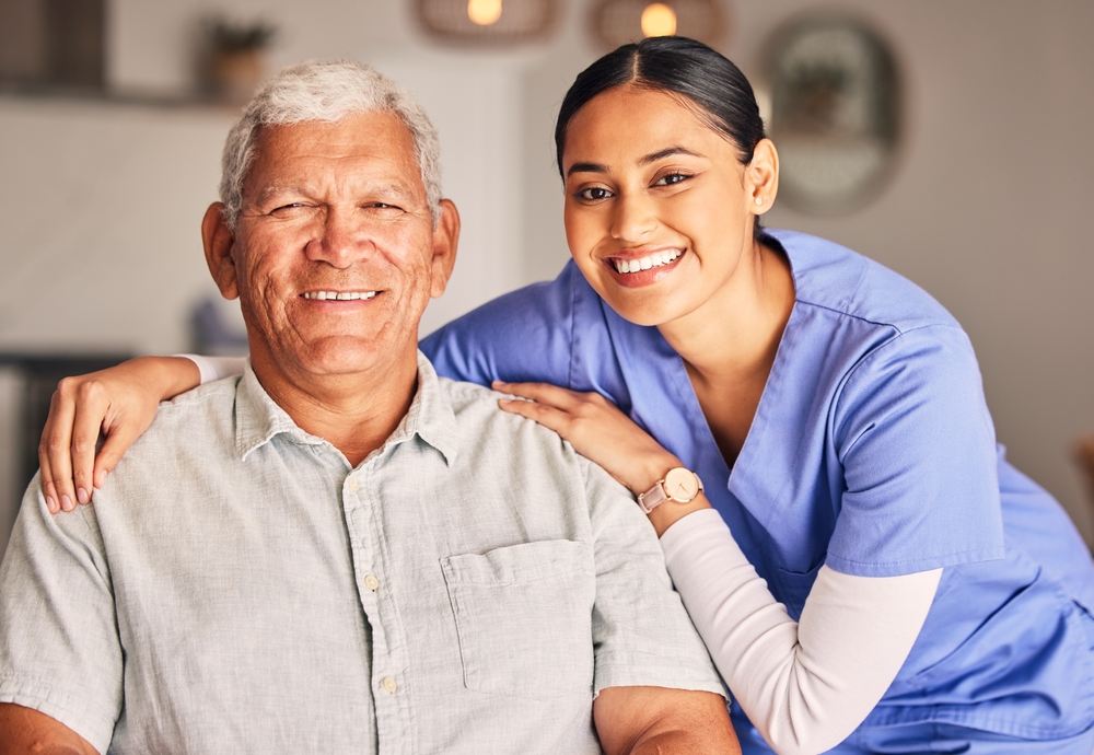 A young Latina health aide in light blue scrubs rests her hands on an elderly male patient's shoulders.