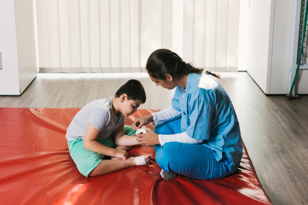 A young South Asian caregiver in blue scrubs works with her special-needs client on a roll of tape while sitting on an orange mat.